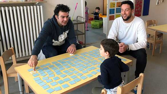 Zwei Väter spielen mit einem Kind Memorie im Kindergarten