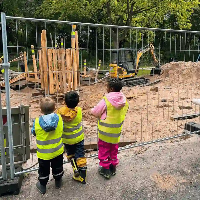 Drei Kinder warten am Bauzaun auf die Fertigstellung des Spielplatzes