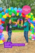 Zentrumsleitung des Kinder- und Familienzentrums Flintacker steht in einem bunten Luftballonbogen und hält eine bunte Ballon-50