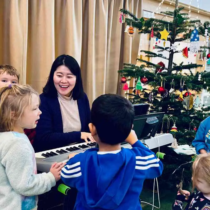P&auml;dagogin sitzt am Keyboard und spielt Musik, umstehende Kinder singen Weihnachtslieder