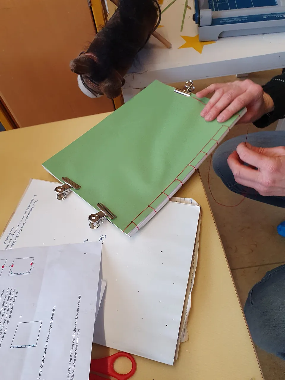 Ein Buch wird hergestellt: Papierstapel und eine Hand die mit Nadel und Faden den R&uuml;cken des Buches n&auml;ht. 