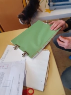 Ein Buch wird hergestellt: Papierstapel und eine Hand die mit Nadel und Faden den Rücken des Buches näht. 