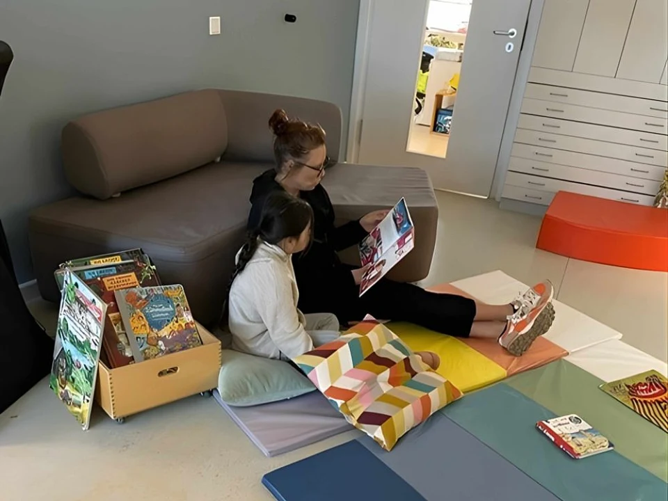 P&auml;dagogin liest einem Kind ein Buch vor. Dabei sitzen beide auf einer Matte auf dem Boden.