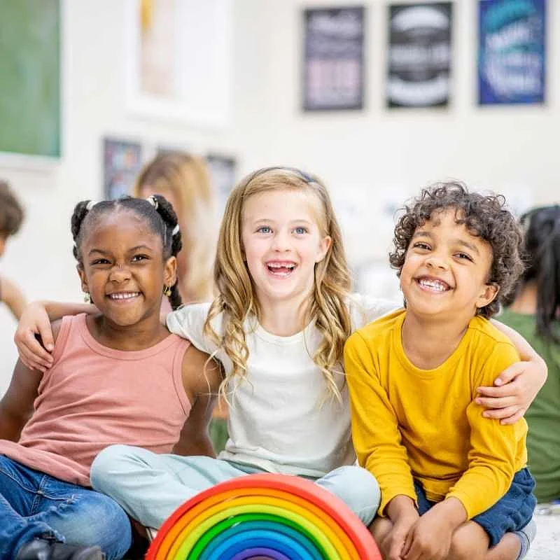 Drei Kinder sitzen nebeneinander auf einem Spielteppich und umarmen sich dabei.