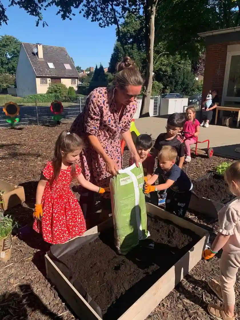 Eine Erzieherin und Kinder bef&uuml;llen das Hochbeet mit frischer Erde