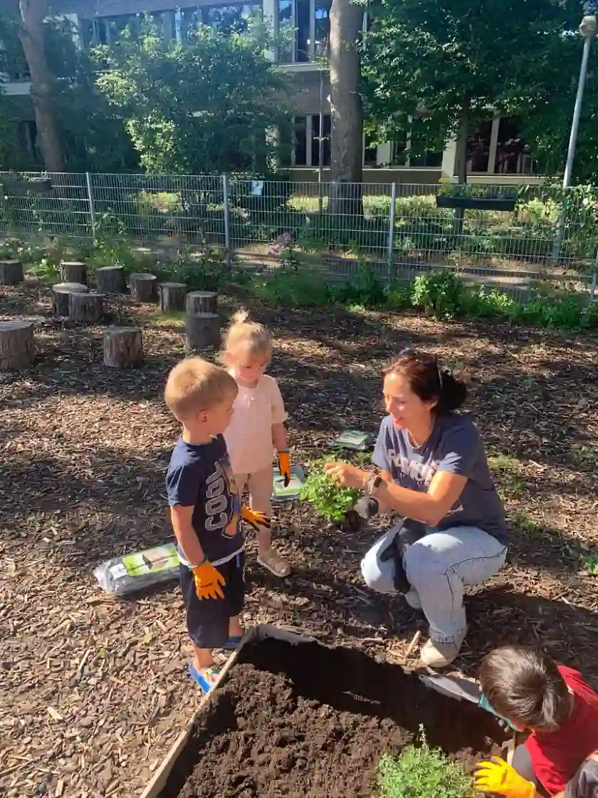 Erziehern zeigt zwei Kindern einen Topf mit frischen Kr&auml;utern f&uuml;r das neue Beet
