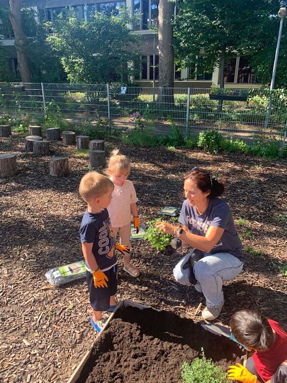 Erziehern zeigt zwei Kindern einen Topf mit frischen Kräutern für das neue Beet