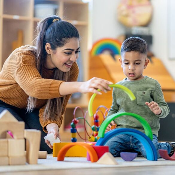 Erzieherin stapelt Holzbögen mit kleinem Jungen. Beide sitzen auf dem Fußboden.